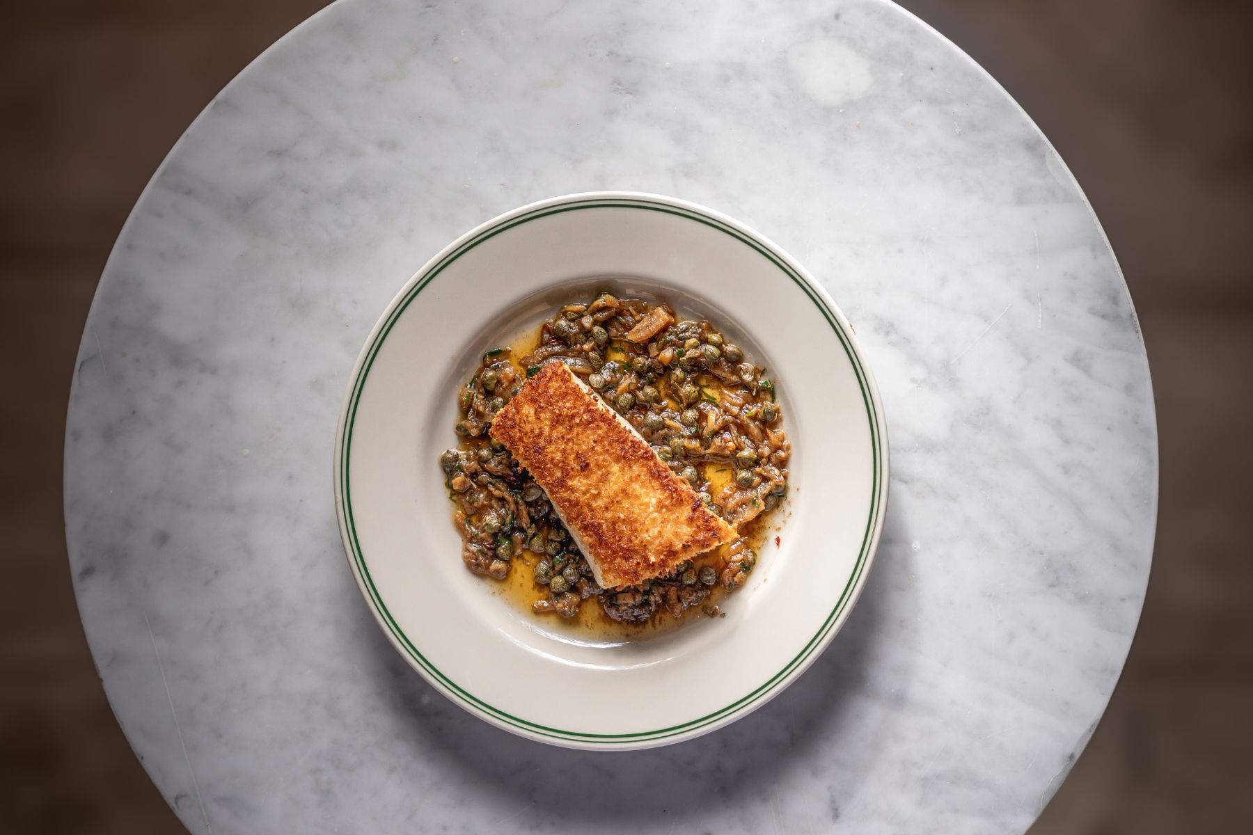 En tallrik med mat, med en bit panerad fisk ovanpå, serverad på en vit tallrik med en grön kant, placerad på ett marmorbord.