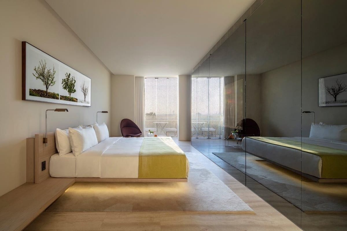 A modern bedroom with a large bed, a sofa, and a chair. There are large windows and a view of the outside.