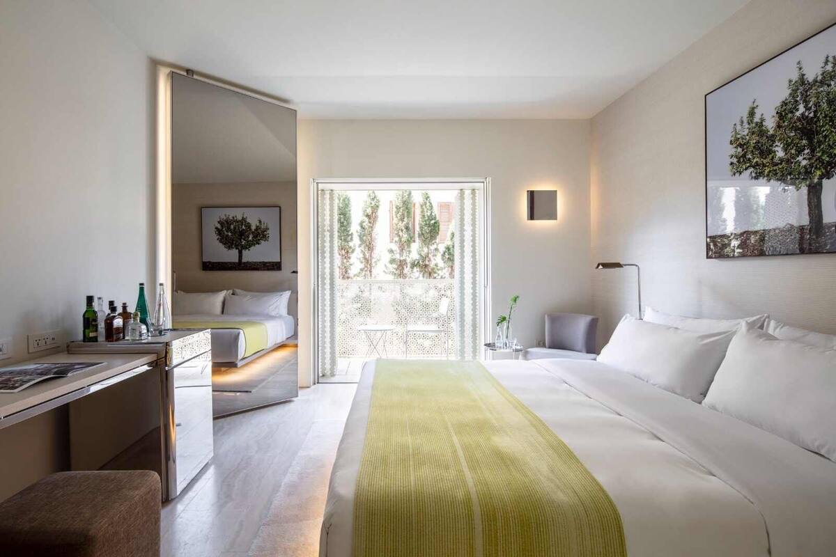 A modern hotel room with a bed, a desk, and a balcony with a view of trees.