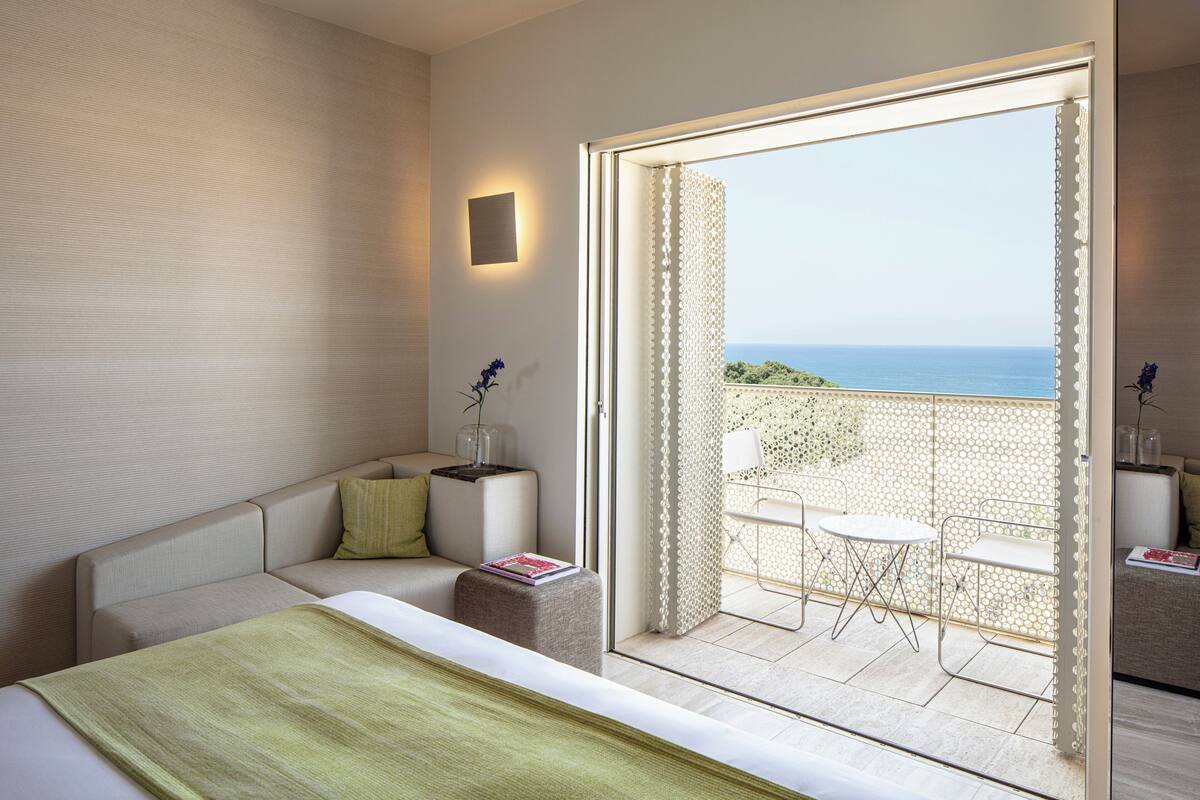 A modern hotel room with a balcony, a sofa, a small table, and a view of the sea.