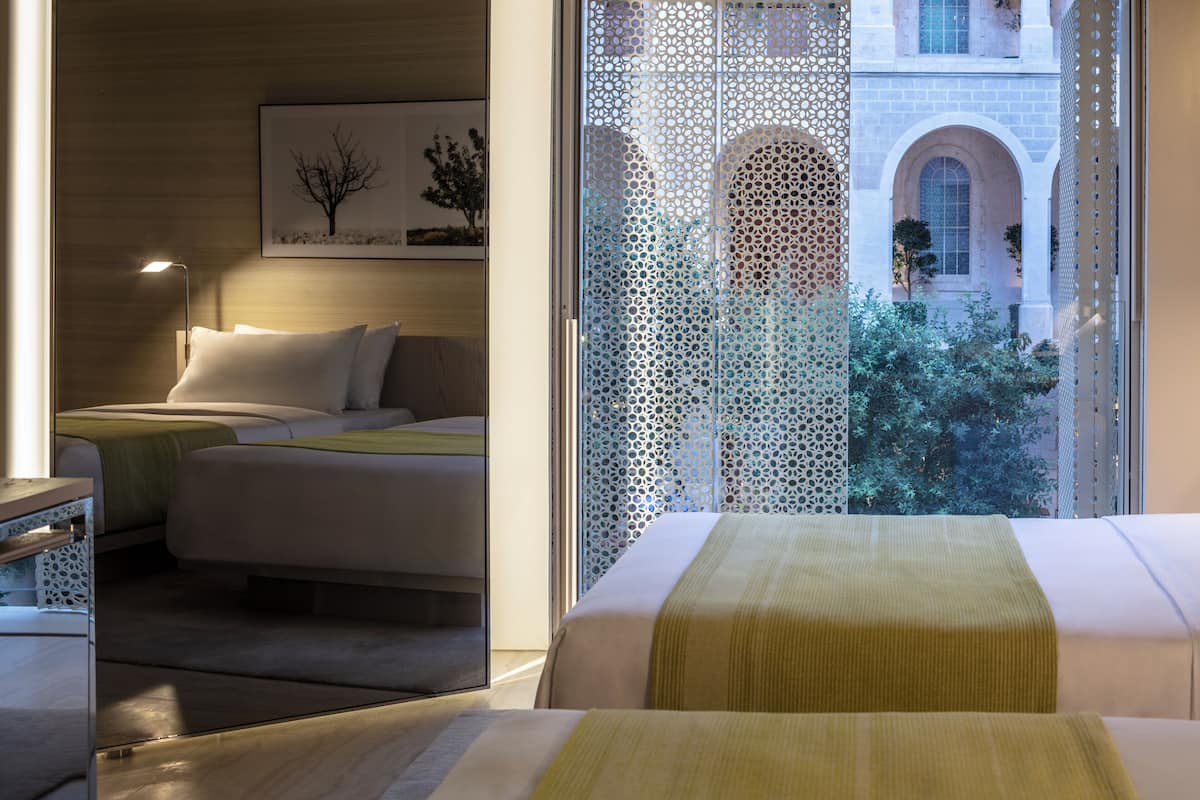 A hotel room with a bed, bedside tables, a lamp, and a view of a building with arches and greenery.