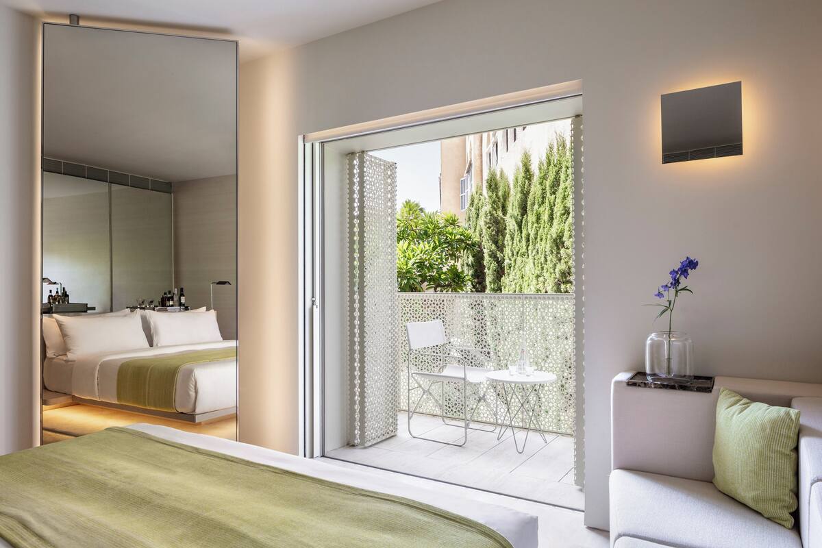 A modern hotel room with a balcony, a bed, a seating area, and a view of greenery.