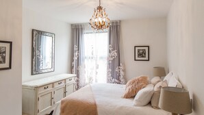 A bedroom with a bed, a dresser, a nightstand, a mirror, and a chandelier. - Provincial Apartment 2 (Nelson)