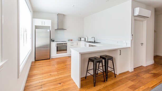 A modern kitchen with white cabinetry, a stainless steel refrigerator, an oven, and a built-in stove. There are two stools at the island counter. - Provincial Apartment 2 (Nelson)