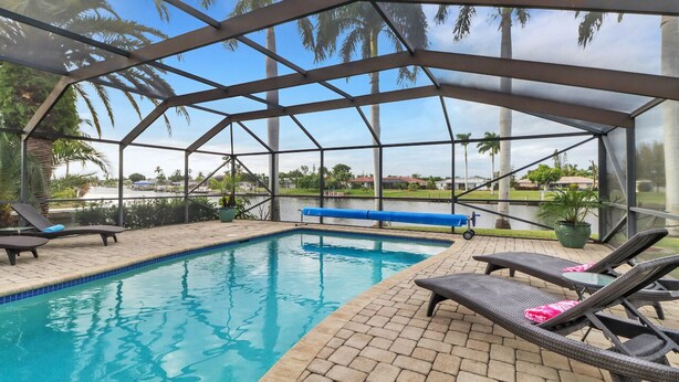A covered swimming pool with lounge chairs and a bench, surrounded by palm trees and a clear sky.