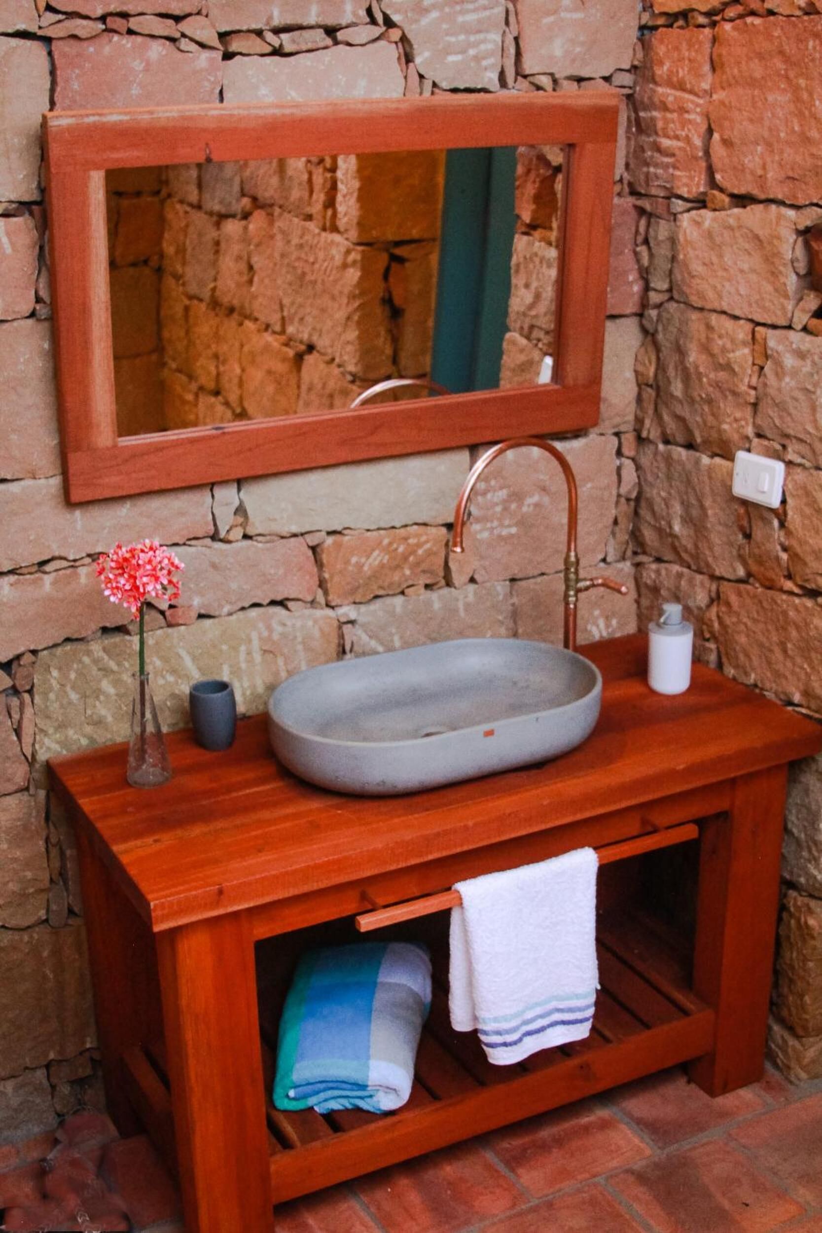 A bathroom with a stone wall, a wooden vanity, a round sink, a mirror, a towel rack, and a small vase with flowers.