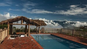 A pool area with a thatched-roof structure, a brick deck, and a view of mountains.