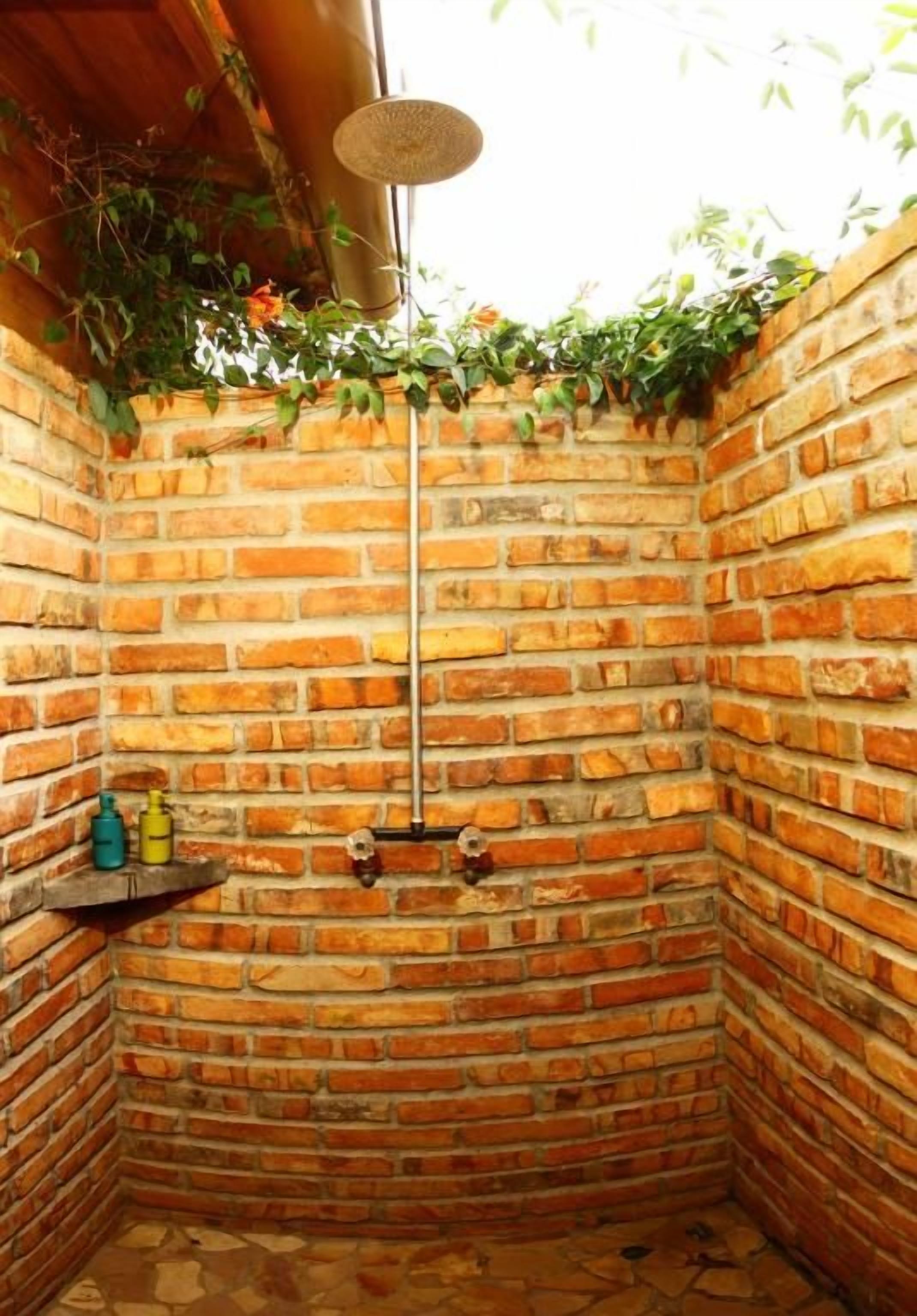 Chuveiro de canto com parede de tijolos, chuveiro pendurado e prateleira com produtos de higiene pessoal.