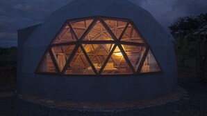 A geodesic dome structure with a triangular window design, a visible interior with a bed and curtains, and a gravel ground surrounding it.