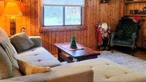A cozy living room with a wooden paneled wall, a sofa, a green armchair, a small table with a Christmas tree, and a window with blinds. - 2 BD/2BA  LOG HOME 1 1/2 MI To SDC 500 YDS TO TABLE ROCK LAKE 15 MIN TO BRANSON (Branson)