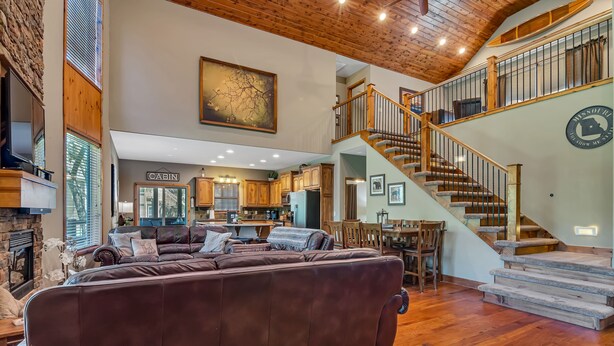 A spacious living area with a large brown leather sofa, wooden flooring, a fireplace, and a staircase leading to an upper level. - Large Side Yard! 2 minutes to Table Rock Lake! (Hollister)