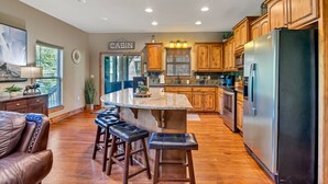 A modern kitchen with wooden cabinets, a large island with stools, a refrigerator, and a dining area with a couch. - Large Side Yard! 2 minutes to Table Rock Lake! (Hollister)