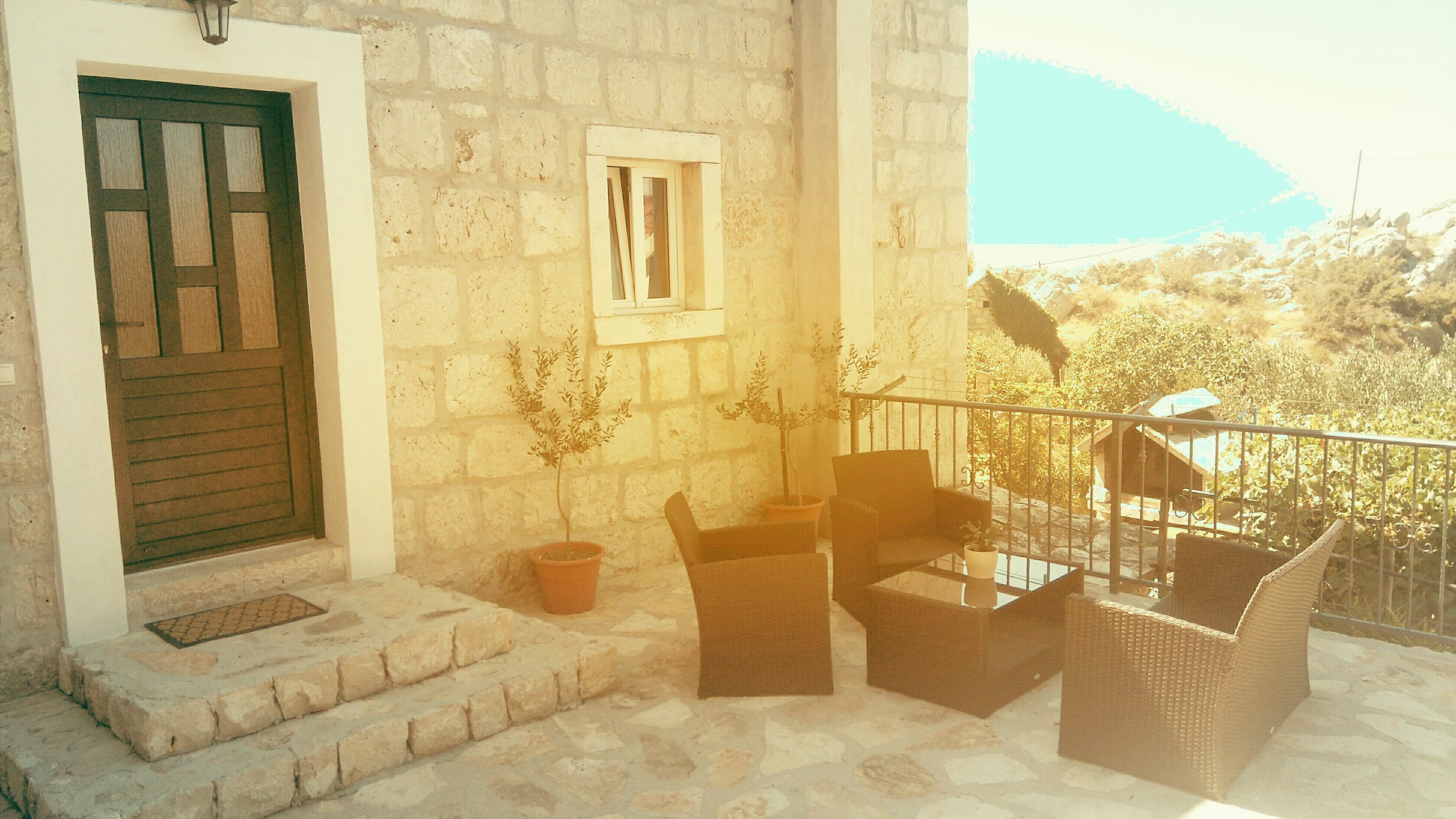 A stone building with a wooden door, a small window, outdoor furniture, and a balcony with a view of the landscape.