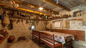 A rustic stone-walled room with a wooden bench and table, shelves with various kitchen utensils, and a hanging lantern. - old stone house Lelas (Omiš)
