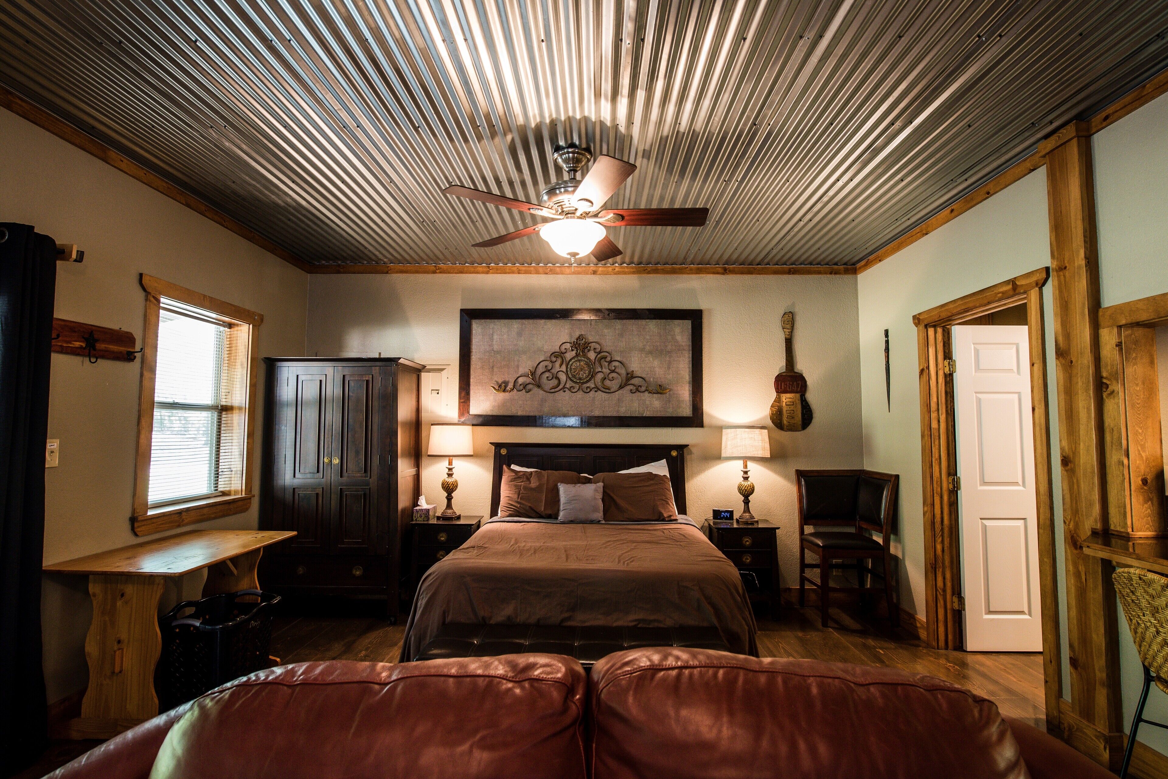 A bedroom with a bed, wooden headboard, two bedside tables with lamps, a ceiling fan, and a large framed artwork above the bed.