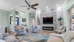 A living room with a fireplace, two armchairs, a sofa, a ceiling fan, and a TV mounted on the wall. - Cecilia 1015 Modern SW Cape Gulf Access Home w/ heated Pool & Spa (Cape Coral)