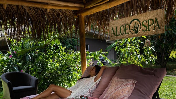 A person relaxing on a bed under a thatched roof with a sign reading "ALOEO SPA".