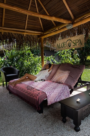 Iemand ontspant op een bed onder een rieten dak, met een bord waarop 'ALOEO SPA' staat.