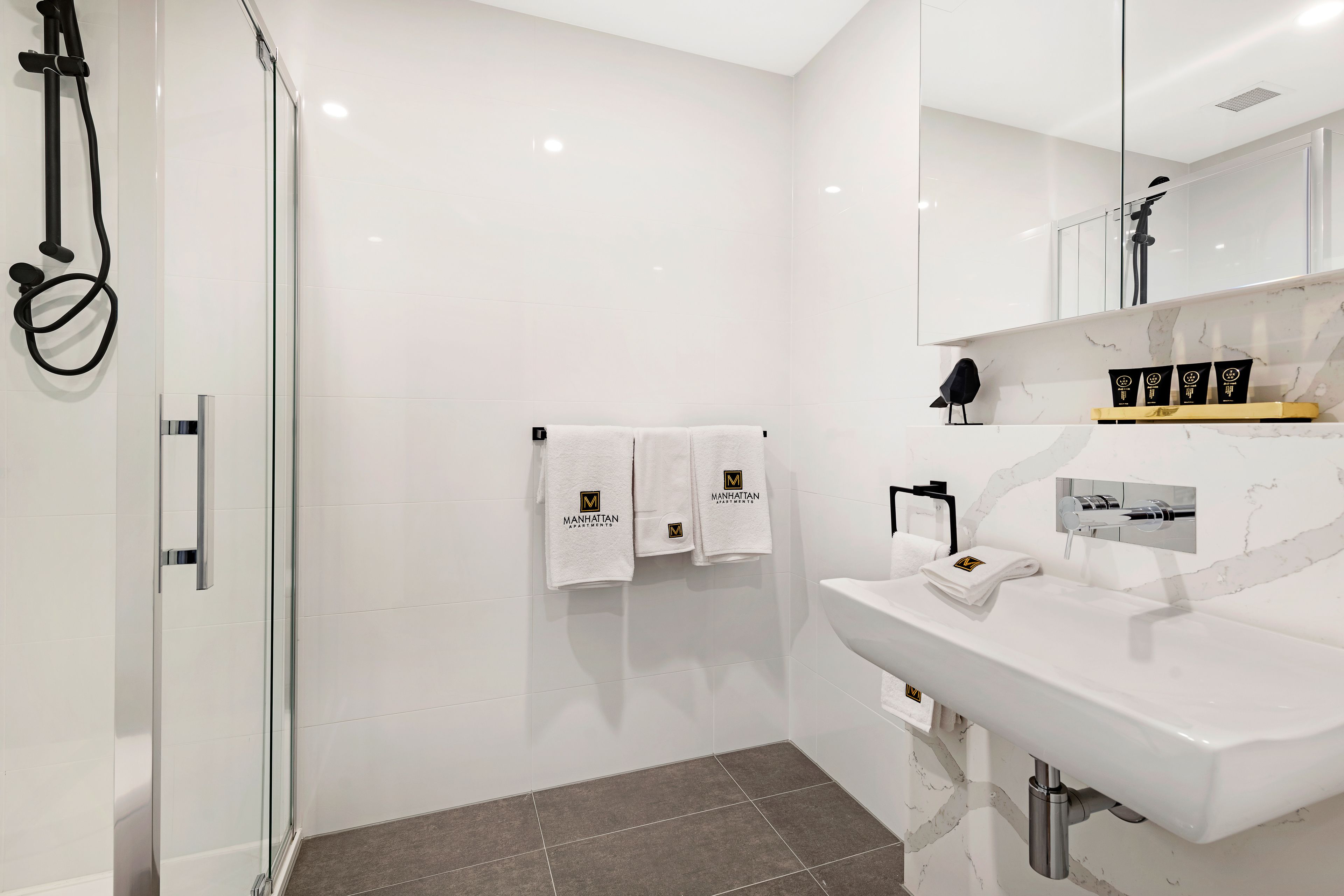 A modern bathroom with a glass-enclosed shower, white tiles, a marble countertop, and towels with a logo.