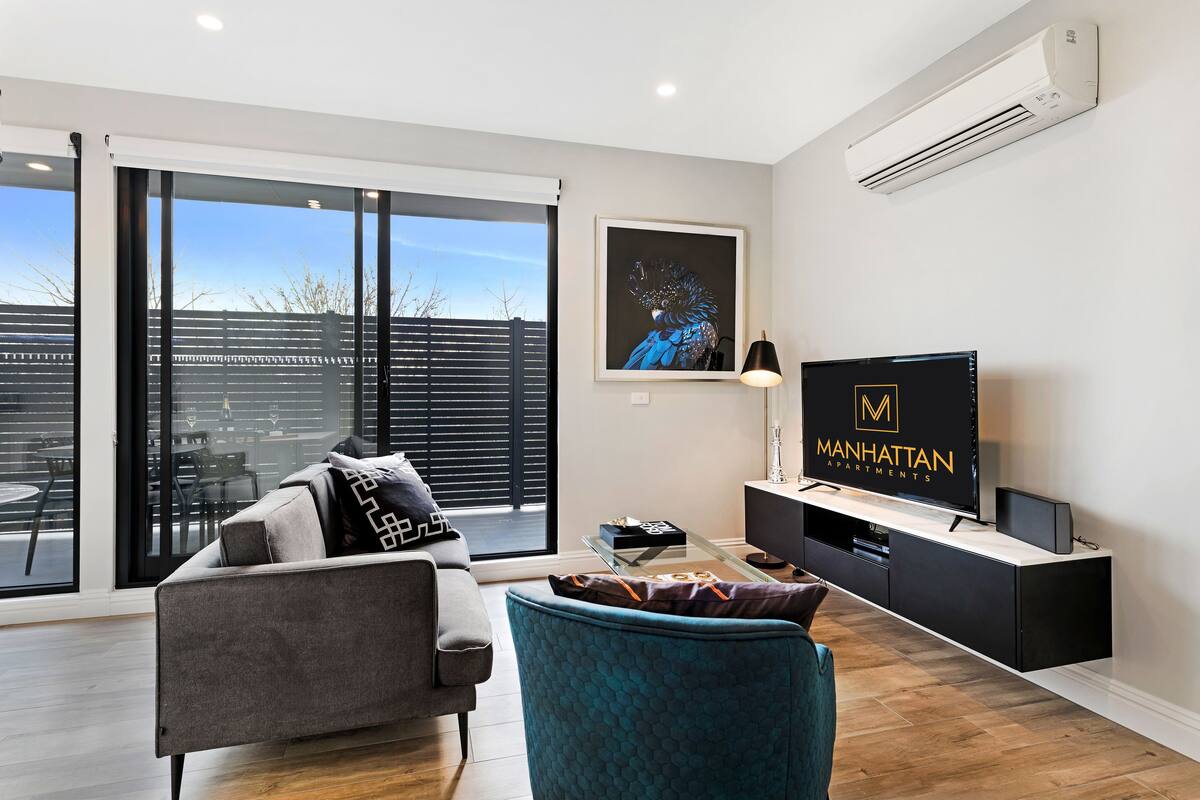 A modern living room with a grey sofa, a teal armchair, a flat-screen TV, and a large window with a view of the outside.