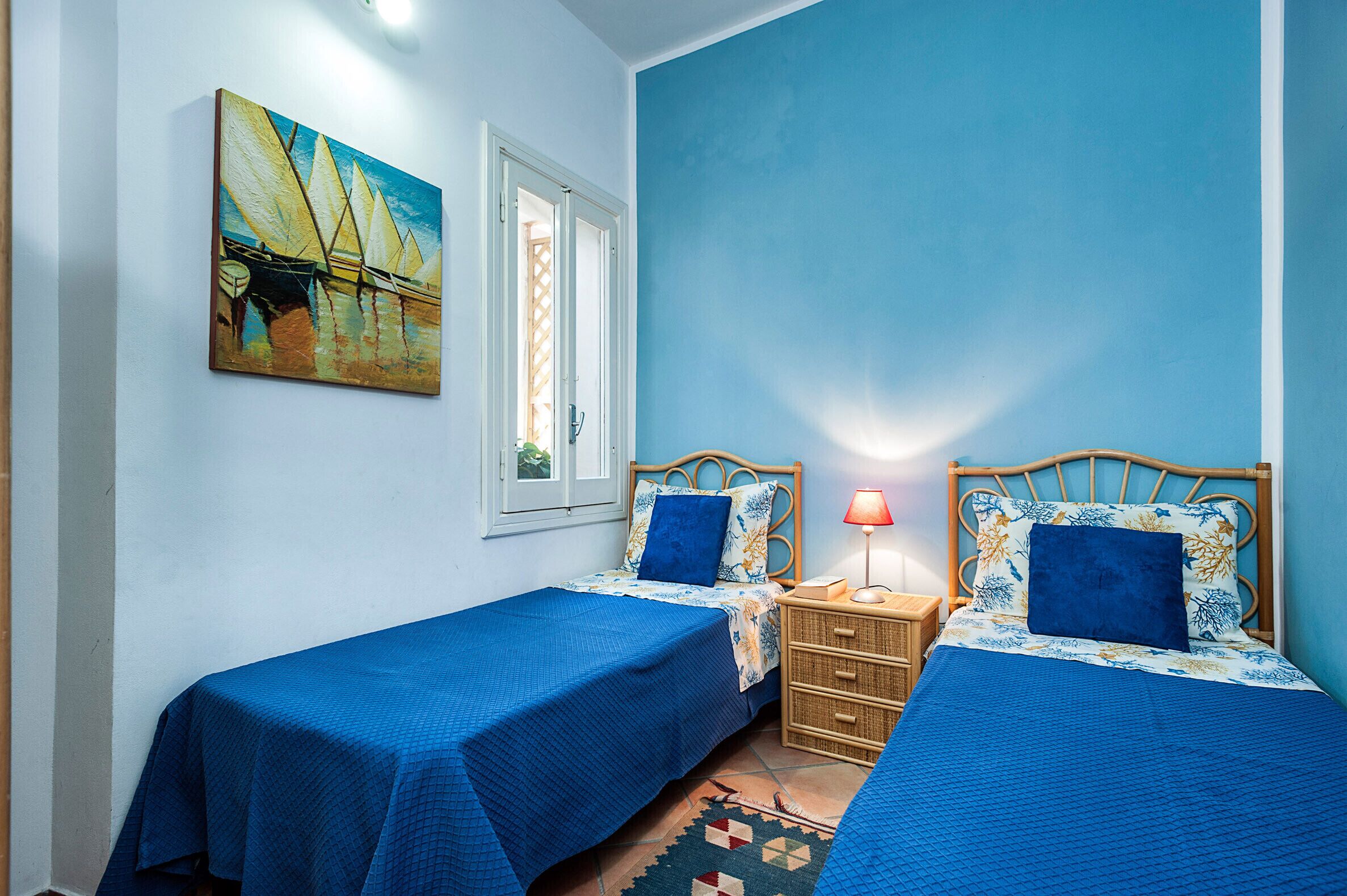 A room with two beds, a painting, a window, and a nightstand with a lamp.