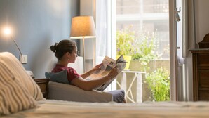 En person sidder på en seng og læser en bog i et rum med et vindue, en lampe og et lille bord med en plante.