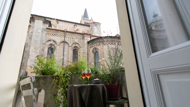 En balkon med et bord, to stole og plantekasser. I baggrunden ses en bygning med en kuppel og buede vinduer.
