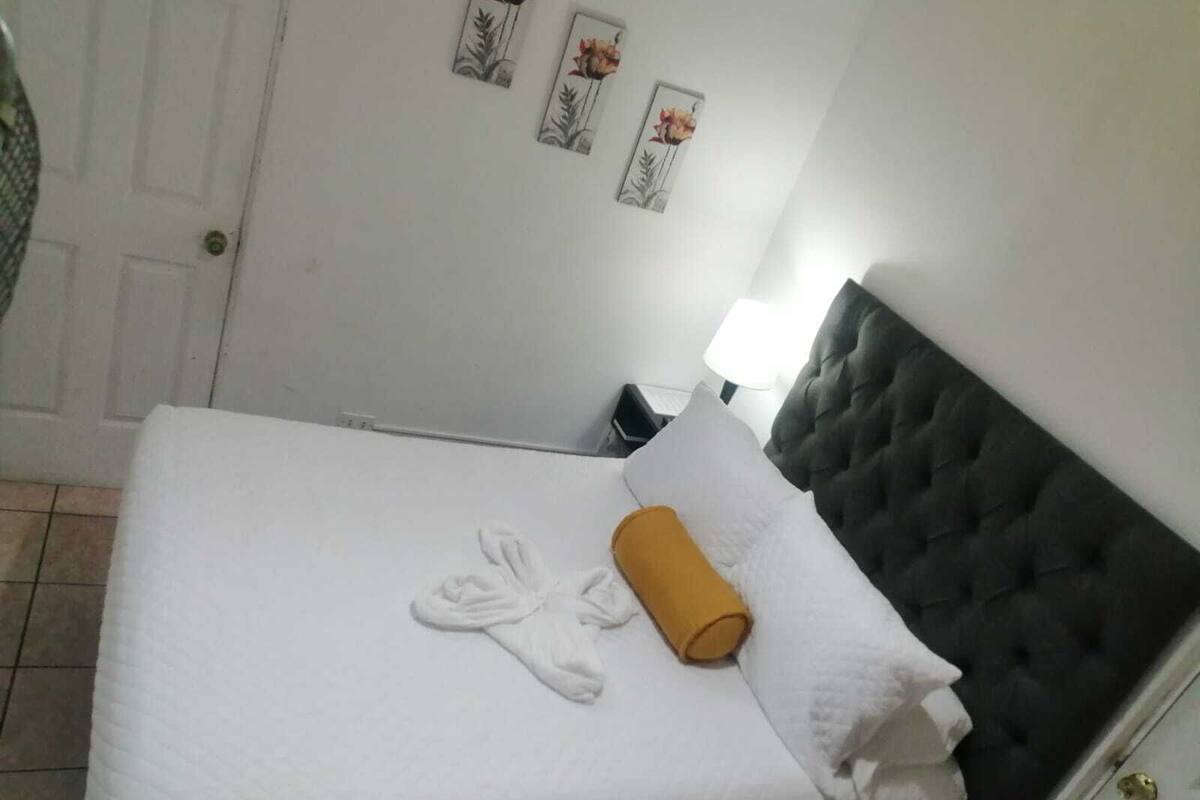 Un lit bien fait, avec du linge de lit blanc et une serviette enroulée, une tête de lit sombre et une lampe sur une table de chevet.