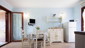 A small kitchen with a dining table, chairs, a TV, a refrigerator, and a wooden cabinet. - A beautiful villaetta overlooking the sea (COSTA REI)