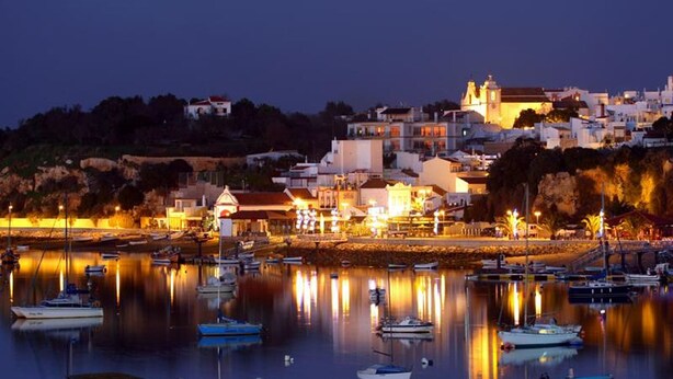 A coastal town at dusk with boats docked in calm waters, a prominent church on a hill, and buildings with warm lighting. - Torralta Alvor Beach 601 (Alvor)