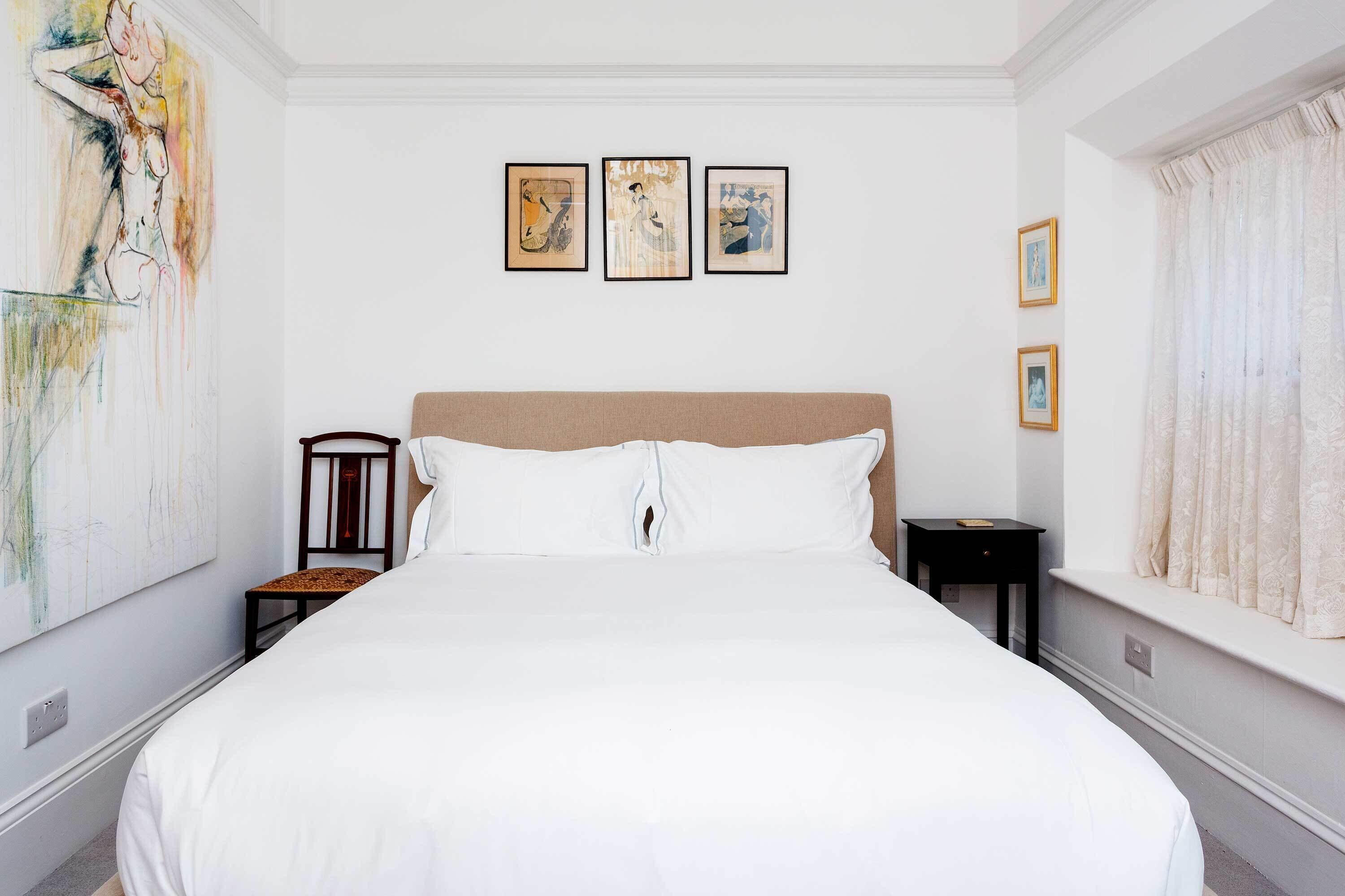 A neatly made bed in a room with a large painting on the wall, a small table, and a window with curtains.