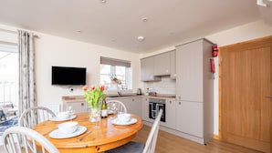 Une cuisine moderne avec des meubles blancs, une table à manger en bois dressée avec de l’assiette blanche, et un téléviseur à écran plat fixé au mur.
