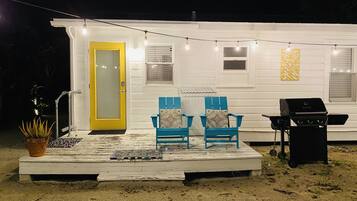 A white house with a porch, two blue chairs, a grill, and string lights.