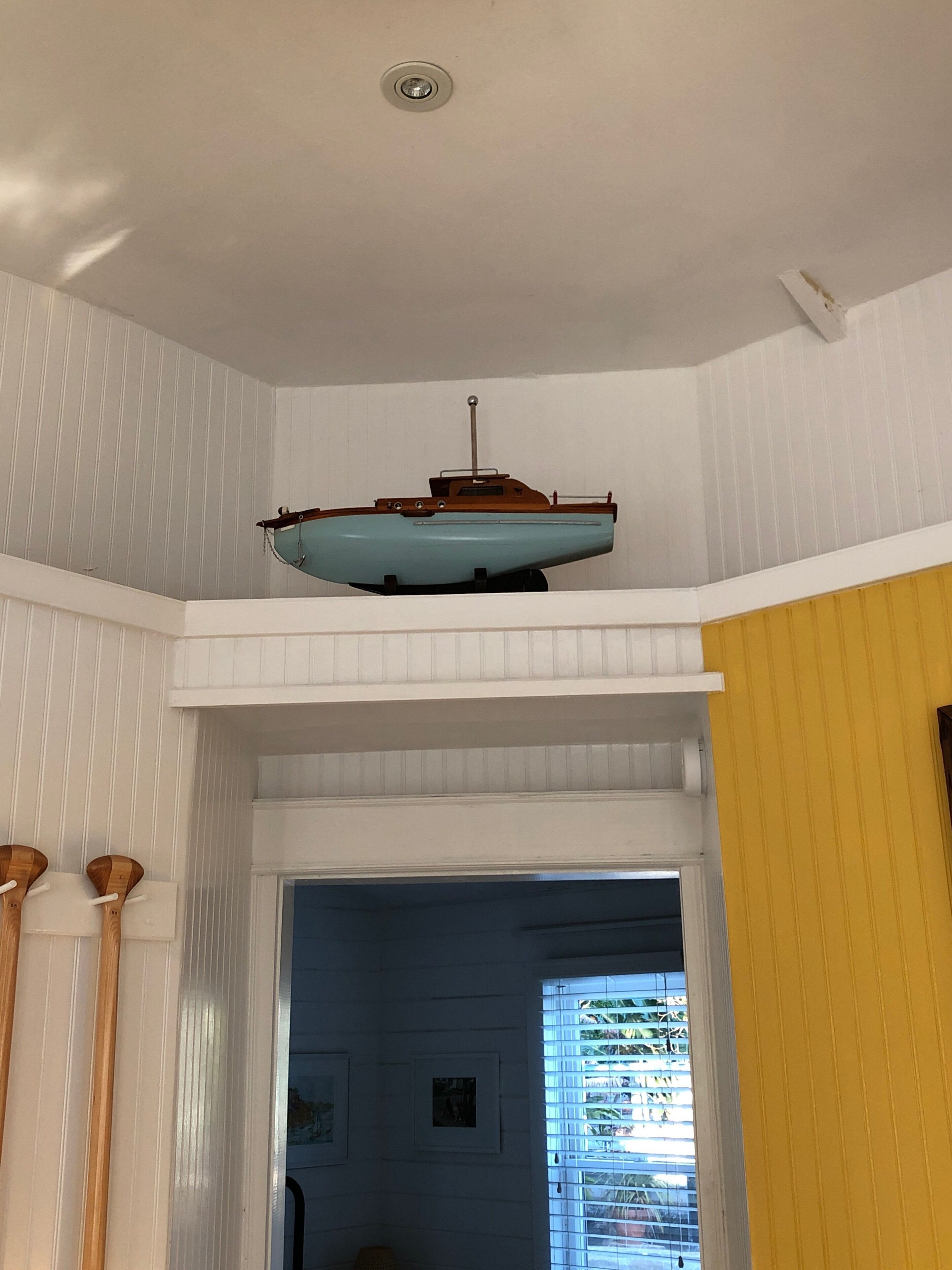 Une pièce avec un modèle de bateau suspendu au plafond, des murs recouverts de panneaux blancs, une porte donnant sur une autre pièce et une fenêtre avec des stores.