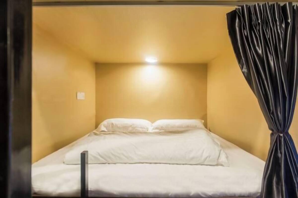 Une petite chambre avec un lit simple, un mur jaune uni, un couvre-lit blanc et un rideau noir sur le côté droit.