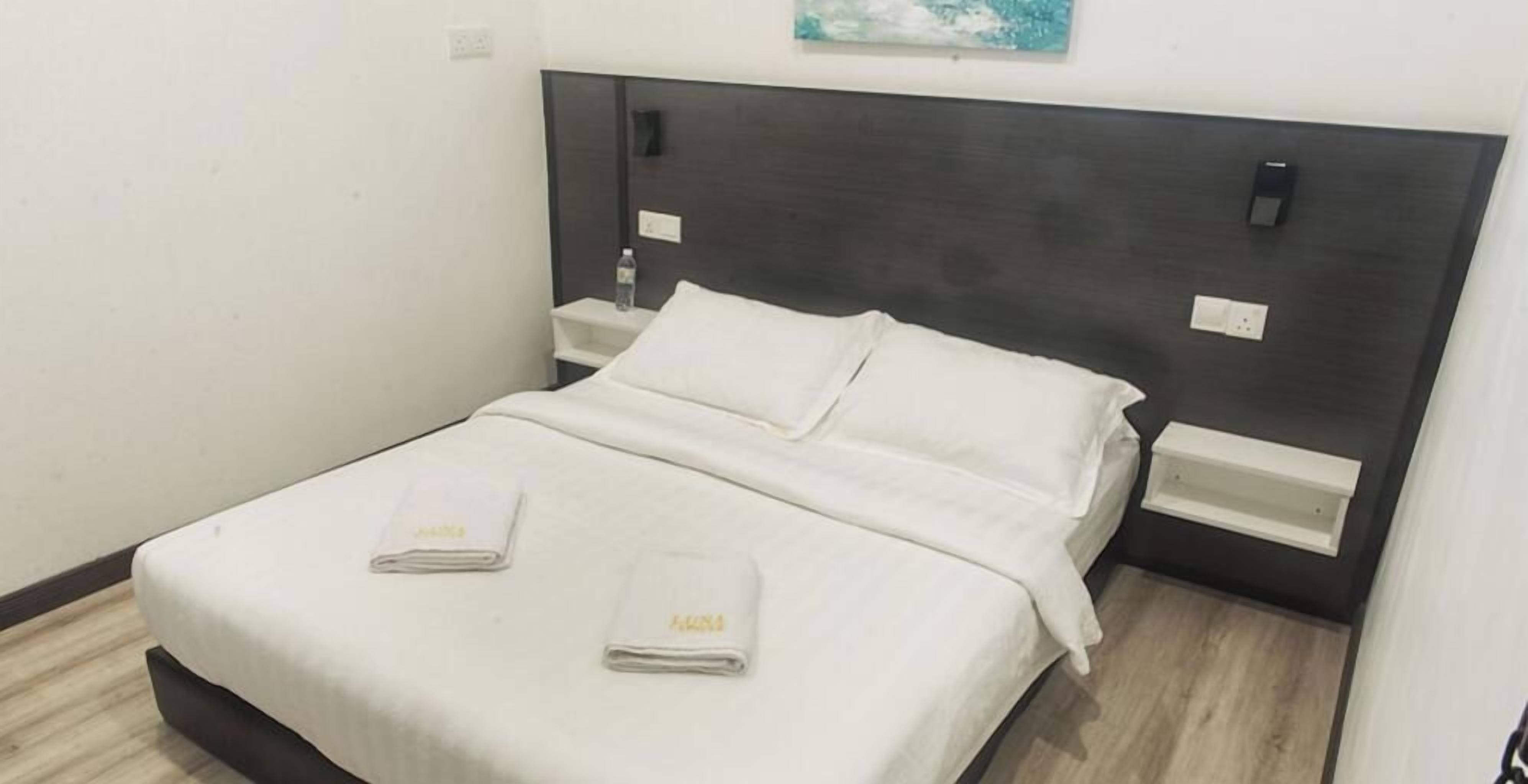A neatly made bed with white linens and two towels, a dark headboard, and a small bedside table with a bottle and a power outlet.