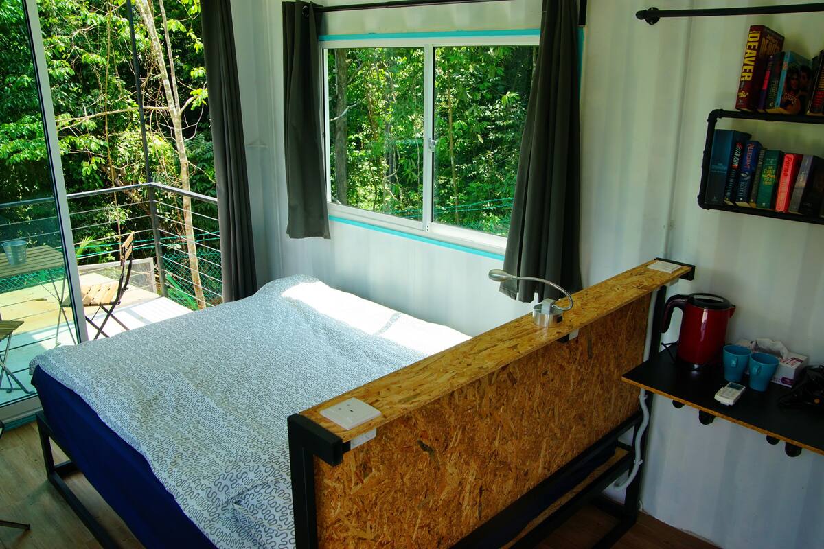 Een slaapkamer met een bed, een raam met gordijnen, een boekenkast en een kleine tafel met een waterkoker en bekers.
