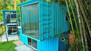 Un edificio modular azul con grandes ventanales y un balcón, rodeado de árboles de bambú.