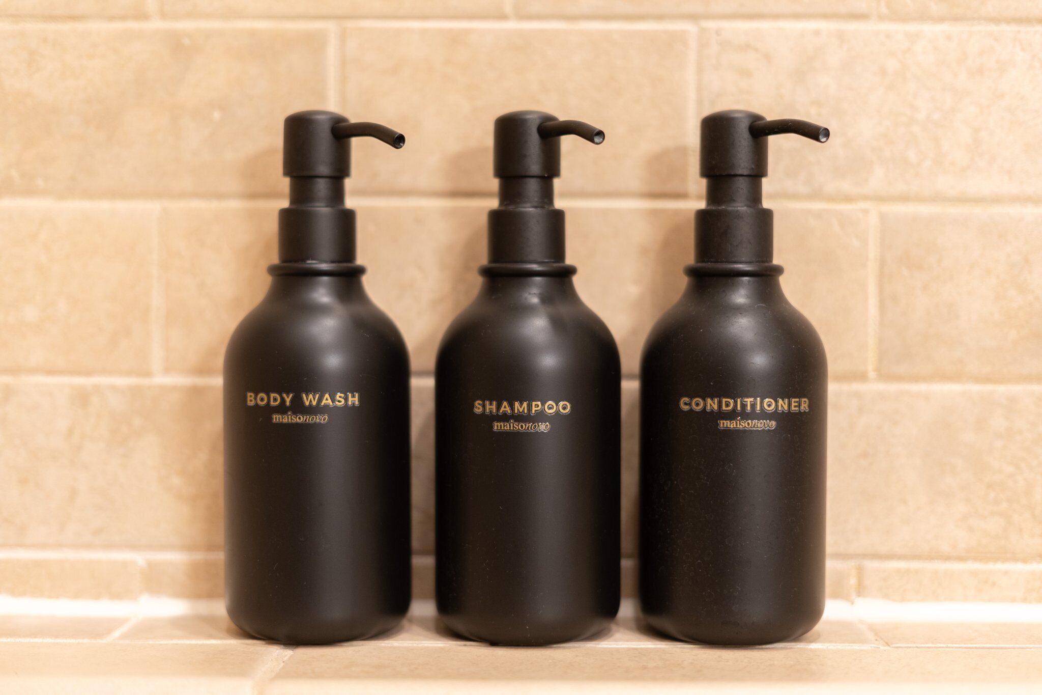 Three black bottles of body wash, shampoo, and conditioner on a tiled surface.