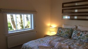 Quarto com cama, roupa de cama floral, janela com vista para árvores, abajur e radiador.