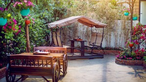 Une table et des chaises en bois sont placées sous un parasol de type balancelle, dans un jardin agrémenté de plantes et de fleurs en pot.