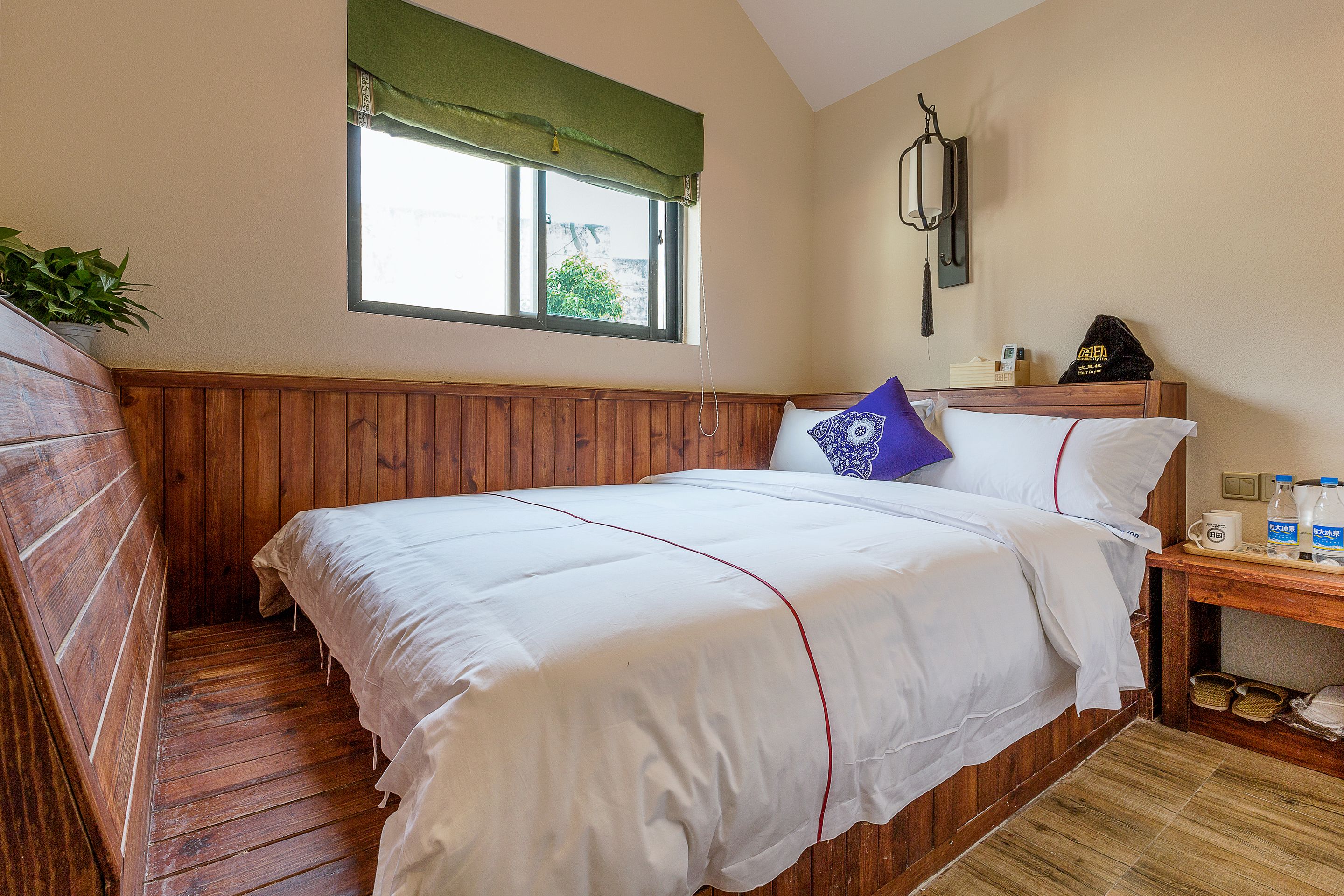A bedroom with a wooden headboard, a single bed with white bedding, a bedside table with a lamp, and a window with a green valance.