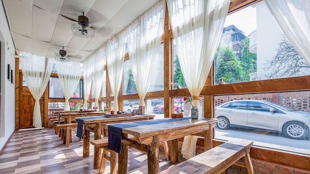 Un restaurant avec des tables et des bancs en bois, de grandes fenêtres ornées de rideaux blancs et une vue sur une rue animée.