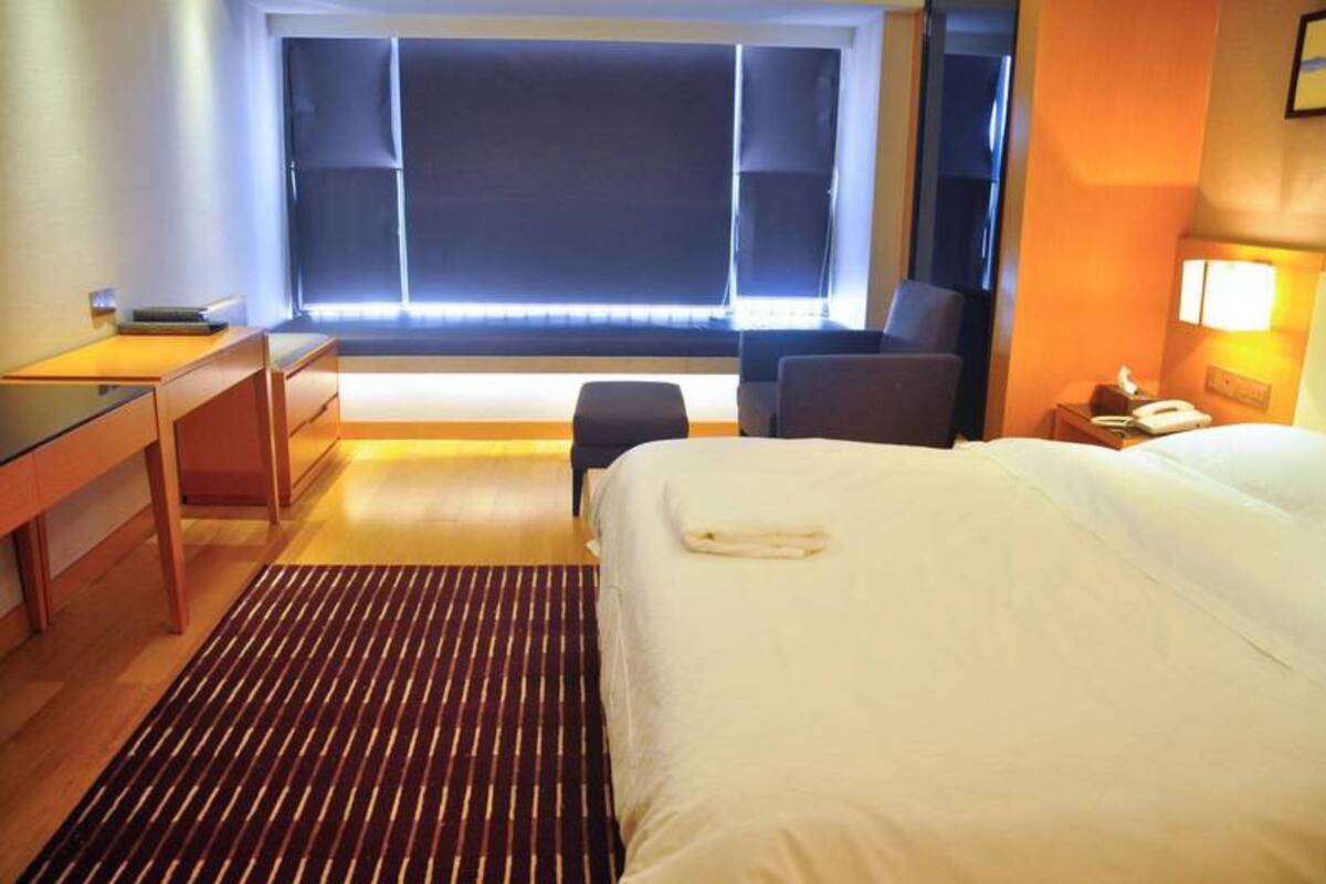 Habitación de hotel con cama, escritorio, silla, mesita, lámpara y ventana con persianas.