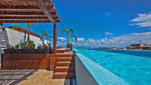 A poolside area with a clear view of the ocean, steps leading to a covered seating area, and a modern building in the background. - MOMA Luxury condo Playa del Carmen (Playa del Carmen)