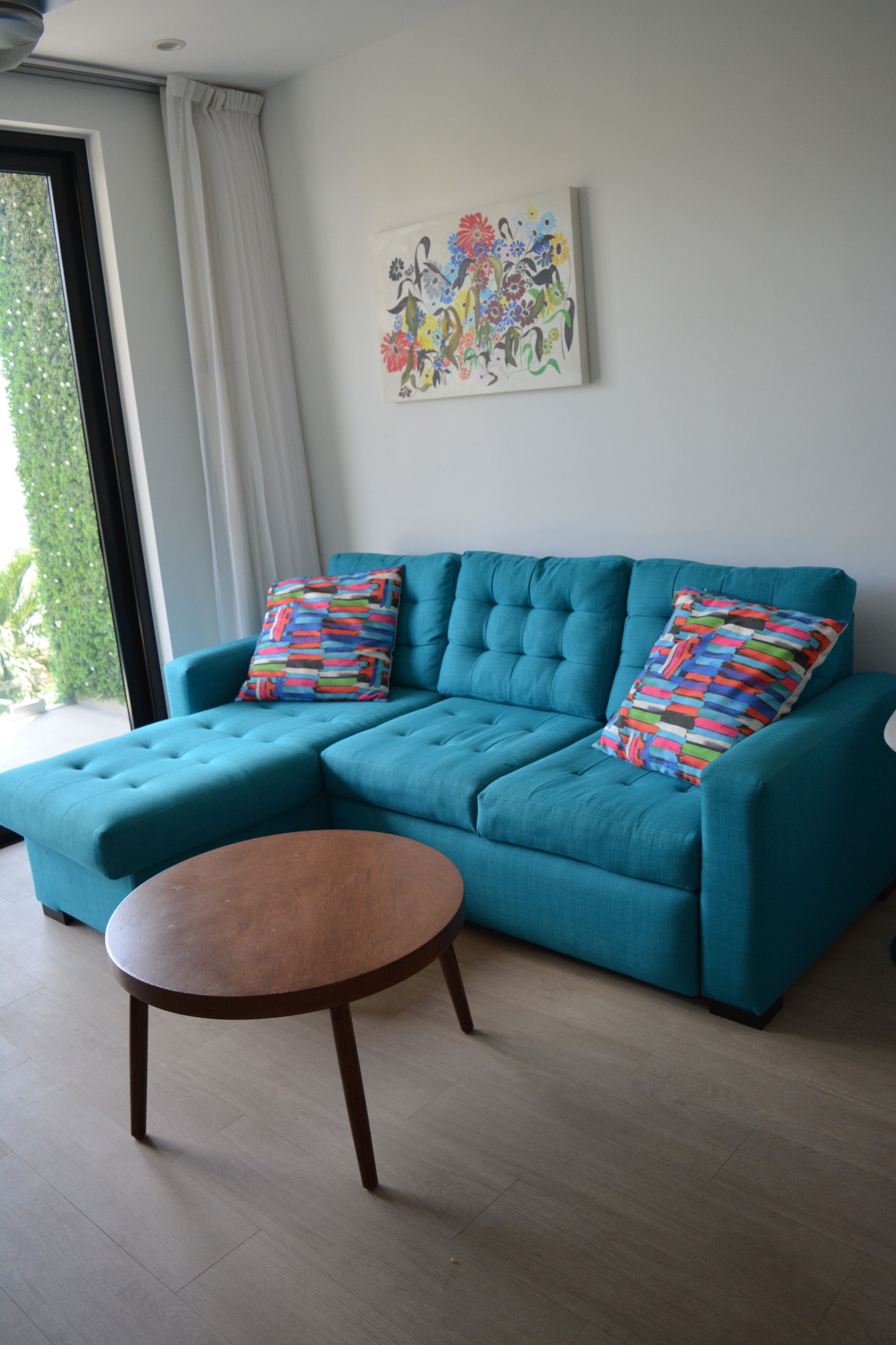 Un canapé de couleur turquoise agrémenté de deux coussins colorés, une table basse en bois et une œuvre d’art florale accrochée au mur.
