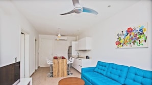 A modern living room with a blue sofa, a wooden coffee table, and a TV on a white stand. There is a ceiling fan, a kitchen area with white cabinets, and a colorful abstract painting on the wall. - MOMA Luxury condo Playa del Carmen (Playa del Carmen)