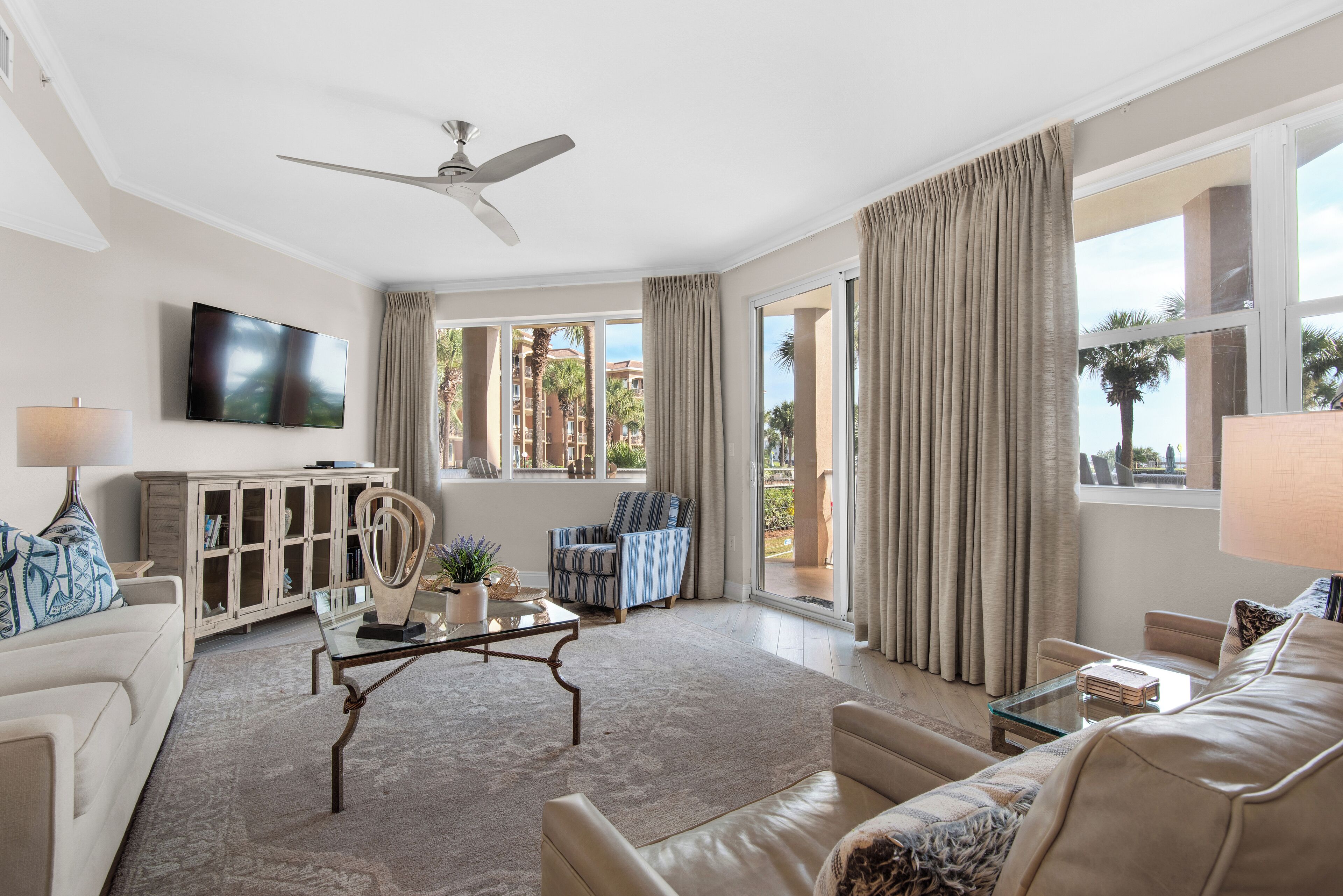 A spacious living room with a ceiling fan, a flat-screen TV, and large windows with curtains.
