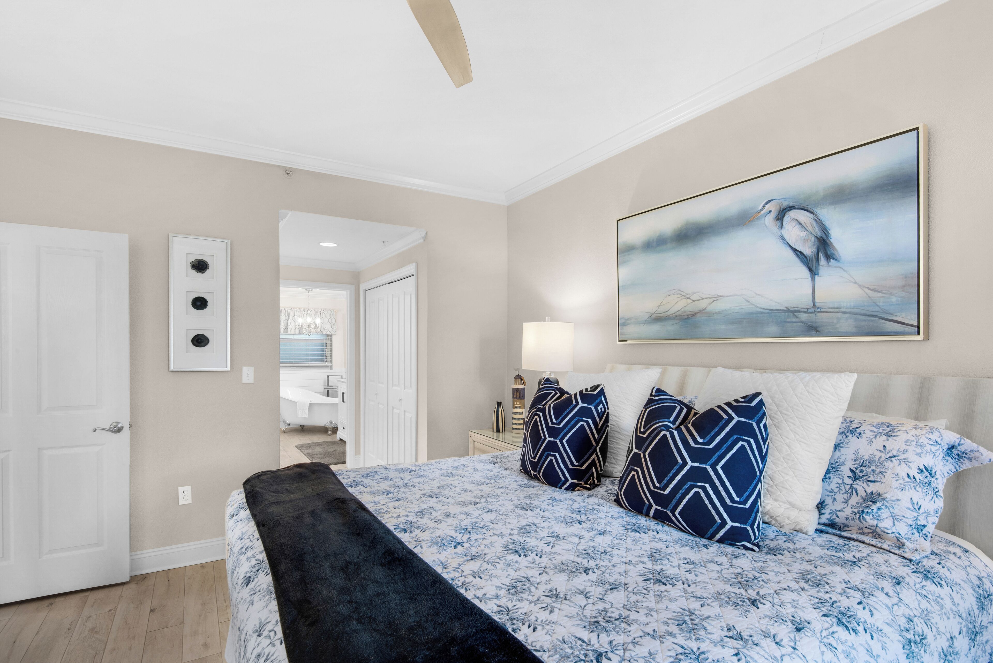 Une chambre avec un grand lit, un ventilateur de plafond, un tableau représentant un oiseau et une salle de bain visible à travers une porte ouverte.