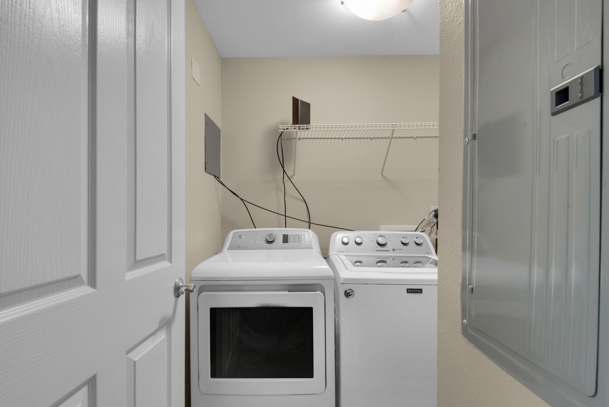 Une buanderie équipée de deux machines à laver et deux sèche-linge superposés, une étagère en métal au-dessus, et une porte sur le côté gauche.